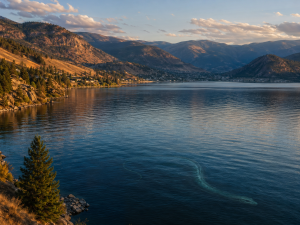 Ein weiter Bergsee bei goldenem Abendlicht mit sanften Wellen, steilen Haengen und einer kaum sichtbaren Wasserwesen-Andeutung unter der Oberflaeche.