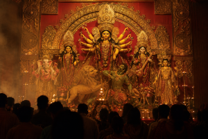 Eine reich geschmueckte Durga-Figur auf einem leuchtenden Festaltar, umgeben von goldenen Ornamenten, Lamplight und der Silhouette von Besuchern.