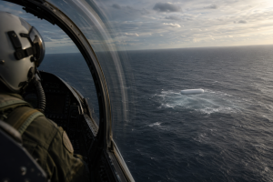 Helles weisses laengliches Objekt ueber einer dunklen Meeresflaeche, beobachtet aus einem Kampfjetcockpit ohne Schrift oder Logos.