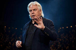 David Icke steht auf einer dunklen Buehne und spricht mit erhobener Hand vor unscharfem Publikum.